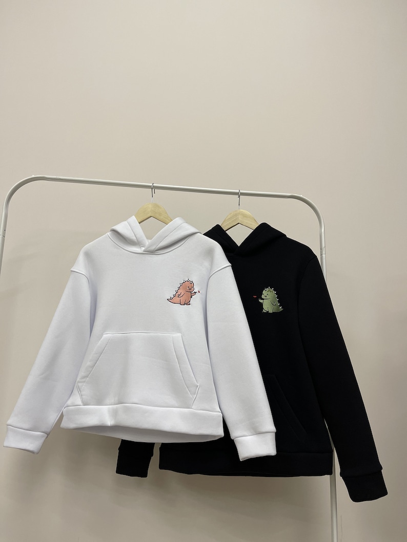 Peut inclure: Deux sweat-shirts &agrave; capuche avec des motifs de dinosaures. L'un est blanc avec un dinosaure rose et l'autre est noir avec un dinosaure vert.