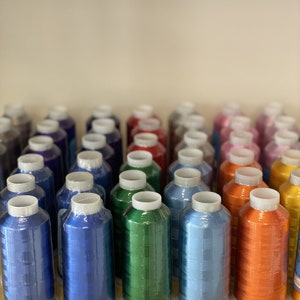 Peut inclure: Une collection de bobines de fil color&eacute;, dispos&eacute;es en rang&eacute;es sur une &eacute;tag&egrave;re. Les bobines sont envelopp&eacute;es de plastique et pr&eacute;sentent une vari&eacute;t&eacute; de couleurs, notamment le bleu, le vert, l'orange et le rouge.