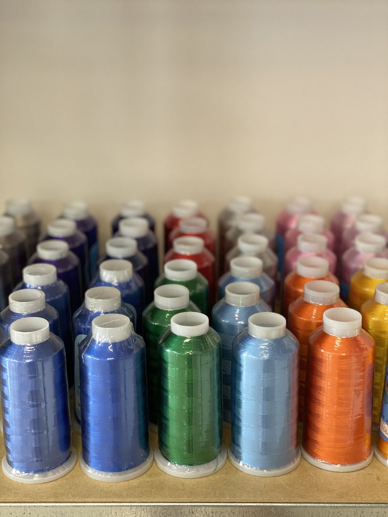 Peut inclure: Une collection de bobines de fil color&eacute;, dispos&eacute;es en rang&eacute;es sur une &eacute;tag&egrave;re. Les bobines sont envelopp&eacute;es de plastique et pr&eacute;sentent une vari&eacute;t&eacute; de couleurs, notamment le bleu, le vert, l'orange et le rouge.