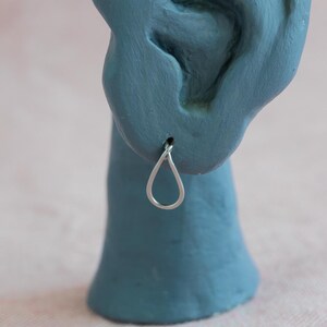Pode incluir: Um brinco prateado em forma de gota é exibido em um modelo de orelha azul. O brinco tem um design simples de gota aberta. Outro brinco com o mesmo design está abaixo. O fundo é rosa claro.