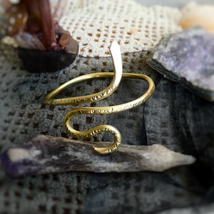 May include: A gold-colored snake arm cuff with a textured design. The snake's head is raised, and the body coils around the arm. The jewelry is displayed on a piece of driftwood, with a blurred background of crystals and fabric.