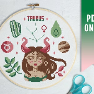 May include: A cross-stitch pattern featuring a Taurus zodiac sign with a female face, green leaves, and a brown planet. The text "TAURUS" is stitched in white on a white background.