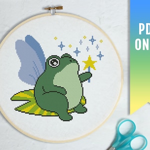 May include: A cross-stitch pattern of a green frog with blue wings and a yellow star wand. The frog is sitting on a yellow and green striped leaf. The background is white with yellow stars.