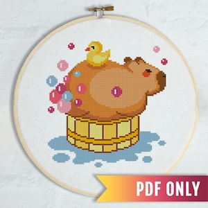 Puede incluir: Un patrón de punto de cruz con un capibara de dibujos animados tomando un baño en una bañera de madera con un pato de goma amarillo. El capibara es marrón y tiene mejillas rosadas. Hay burbujas azules y rosas alrededor del capibara.