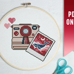 May include: A cross stitch pattern featuring a pink and red camera with a photo of a whale in a pink and blue sky. The pattern also includes two pink hearts.