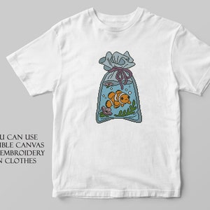 May include: White t-shirt with an embroidered design of a clownfish in a bag of water. The text "YOU CAN USE SOLUBLE CANVAS FOR EMBROIDERY ON CLOTHES" is printed below the design.