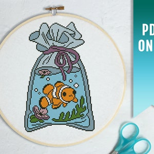 May include: A cross-stitch pattern of a clownfish in a bag with a purple ribbon. The bag is filled with water and has bubbles and seaweed. The pattern is in a hoop.
