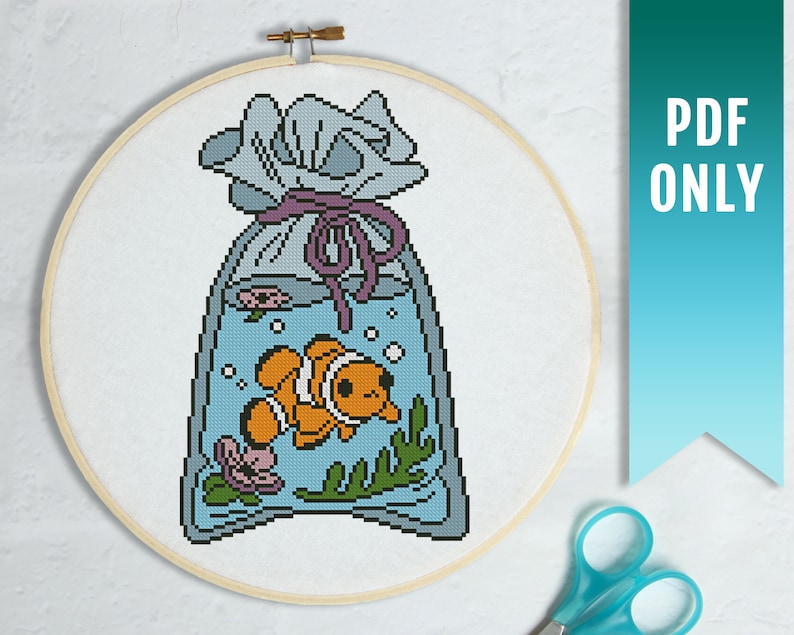 May include: A cross-stitch pattern of a clownfish in a bag with a purple ribbon. The bag is filled with water and has bubbles and seaweed. The pattern is in a hoop.