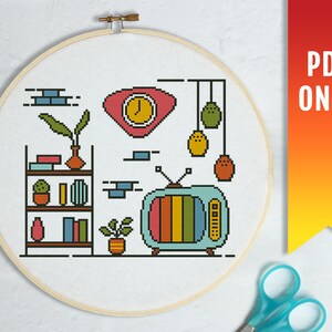 Könnte beinhalten: Kreuzstichmuster mit einer Retro-Wohnzimmer-Szene mit einem Bücherregal, einer Topfpflanze, einer Uhr, einem Fernseher und hängenden Ornamenten. Das Muster befindet sich in einem runden Rahmen mit weißem Hintergrund.