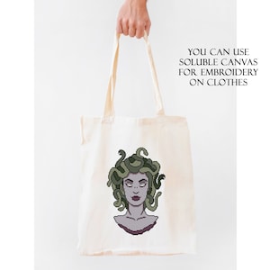 May include: A white tote bag with a green and purple illustration of Medusa, a mythical Greek figure with snakes for hair. The text "YOU CAN USE SOLUBLE CANVAS FOR EMBROIDERY ON CLOTHES" is printed on the bag.