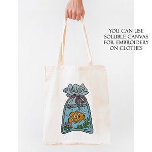 May include: A white tote bag with a blue and orange cartoon fish in a bag with a purple ribbon. The text "YOU CAN USE SOLUBLE CANVAS FOR EMBROIDERY ON CLOTHES" is printed on the bag.