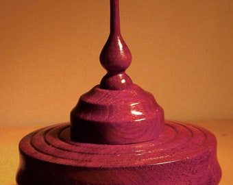 Op de afbeelding: Een houten doos met een paars-rode afwerking en een decoratief houten ornament bovenop. De doos staat op een lichtbruin oppervlak.