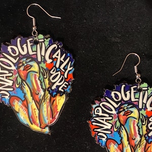 May include: A pair of colorful earrings with a woman's face and the words "Unapologetically Dope" on them.
