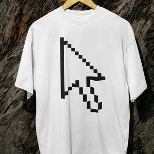 May include: White t-shirt featuring a large black pixelated computer cursor graphic. The design is centered on the front of the shirt. The shirt is hanging on a wooden hanger against a textured brown background.