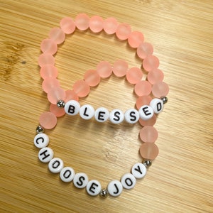 May include: Two pink beaded bracelets with white letter beads spelling out "Blessed" and "Choose Joy".