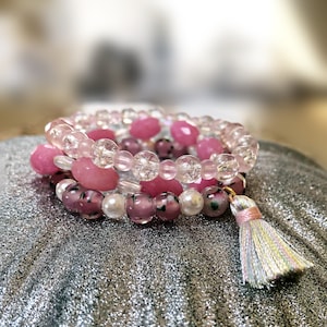 May include: A set of four pink and clear beaded bracelets with a pink and white tassel charm. The bracelets are stacked on a silver background.