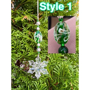 May include: A clear snowflake ornament with a string of green and silver beads, and a green and white glass bead. The text "Style 1" is in the upper left corner. The ornament hangs against a backdrop of green foliage.