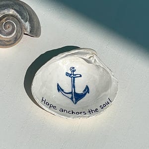 May include: A white seashell trinket dish with a blue anchor design and the text "Hope anchors the soul." A gray and white seashell is in the background. Nautical-themed decor.