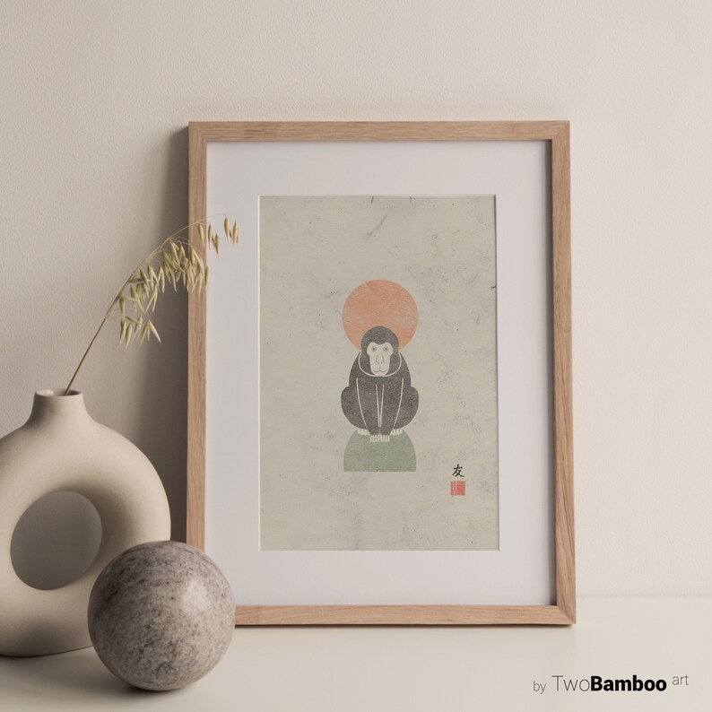 May include: A framed print of a black and white illustration of a monkey sitting on a green rock with a red sun behind it. The print has the Japanese character for "monkey" in red ink in the bottom right corner. The print is framed in a light wood frame.