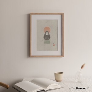 May include: A framed print of a black and white illustration of a monkey sitting on a green platform with a red sun behind it. The print is on a textured paper with a red stamp in the bottom right corner. The frame is light brown wood.