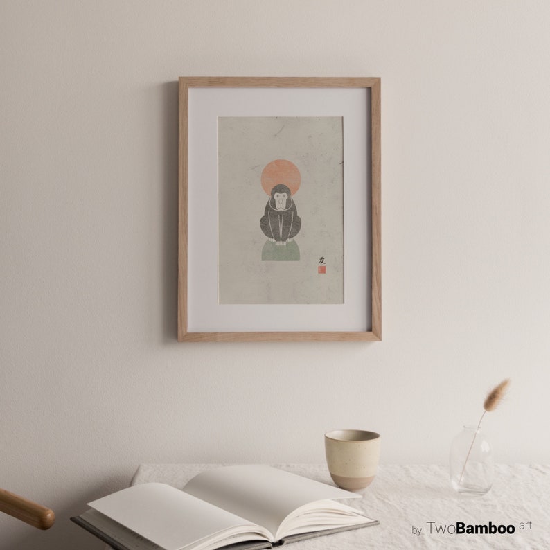 May include: A framed print of a black and white illustration of a monkey sitting on a green platform with a red sun behind it. The print is on a textured paper with a red stamp in the bottom right corner. The frame is light brown wood.