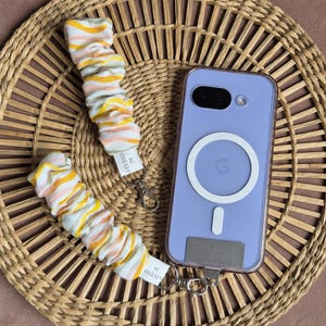 May include: A lavender phone case with a white circular design and a silver clasp. Two fabric wrist straps with yellow, orange, and white stripes are attached to the case. The straps have the text "STUDIO 39".