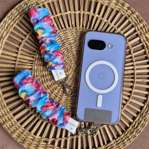May include: A lavender phone case with a white circular design and a matching colorful wrist strap. The strap features a watercolor pattern in shades of blue, red, and yellow. The strap has the text "Studio 39" on a white tag.