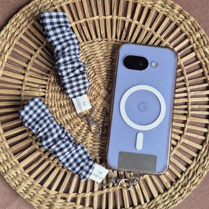 May include: A lavender phone case with a white circular design and a matching black and white gingham wrist strap. The strap has a silver clasp and a tag that reads "STUDIO 39". The phone and strap are on a woven basket.