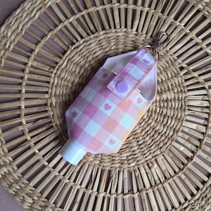 Könnte beinhalten: Ein rosa-weiß karierter Stoff-Handdesinfektionsmittelhalter mit einem silberfarbenen Clip. Der Halter hat ein rosa Herzmuster und einen hellvioletten Druckknopfverschluss. Der Halter befindet sich auf einem gewebten, kreisförmigen Platzset.