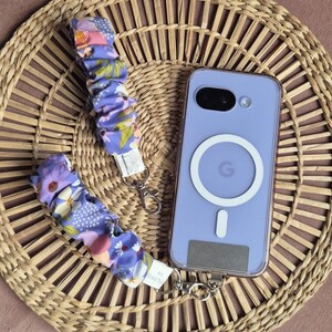 May include: A lavender phone case with a white circular design and a matching floral wrist strap. The strap features a purple, pink, and yellow floral pattern. The phone and strap are on a woven wicker mat.