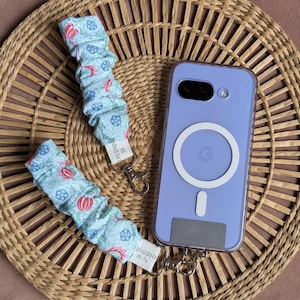 May include: A light blue floral phone wrist strap with red and blue accents, next to a lavender phone case with a white circular design. The strap has a silver clasp and a white tag with black text. The items are on a woven basket.