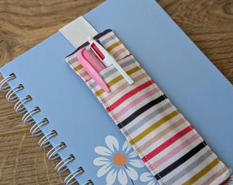 May include: A light blue spiral-bound notebook with a daisy pattern, featuring a striped pen holder with a pink pen and a white pen. The pen holder has a striped pattern with red, black, yellow, and gray stripes.