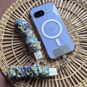 May include: A clear phone case with a purple phone inside, a floral wrist strap, and a matching floral phone charm. The wrist strap and charm have a white tag that reads "STUDIO 39". The phone is resting on a woven, circular placemat.
