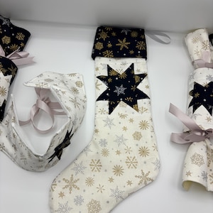 Könnte beinhalten: Weihnachtsstrumpf und festliche Dekorationen. Der Strumpf ist cremefarben mit goldenen und silbernen Schneeflocken und einem schwarzen Stern. Die Manschette ist schwarz mit goldenen Schneeflocken. Weitere Artikel sind eine Gesichtsbedeckung und Knallbonbons, alle mit einem ähnlichen Schneeflockenmuster.