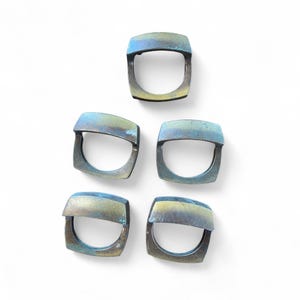 May include: Five square-shaped rings with a metallic finish. The rings have a D-shaped opening and a slightly curved top. The color of the rings is a mix of blue, green, and gold, with a distressed appearance. The rings are displayed on a white background.