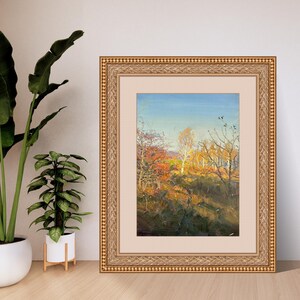 May include: An oil painting in a gold frame depicts an autumn landscape. The artwork showcases trees with vibrant red, orange, and yellow foliage under a clear blue sky. The painting is framed with a gold ornate border.