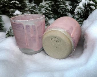 May include: Two handmade ceramic cups with a pink glaze and white drips. One cup is upright, the other is on its side. The cups have a gray base and are set in the snow, with snow-covered evergreen trees in the background.