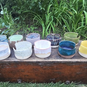 Puede incluir: Una colección de nueve cuencos de cerámica para lana en varios colores, incluyendo azul, verde, amarillo, blanco y rosa. Cada cuenco presenta una forma redondeada y lisa con un corte en forma de U en el lateral, diseñado para sostener la lana al tejer.
