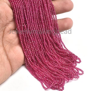 May include: A hand holds a bundle of small, faceted, deep pink beads. The beads are strung together, forming multiple strands. The beads are tightly packed together, creating a solid mass of color. The background is white.