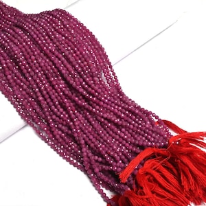 May include: A long strand of small, faceted, deep magenta-colored beads arranged in multiple parallel rows. The beads are strung together and secured at one end with a bright red fabric tassel, creating a uniform and textured appearance.