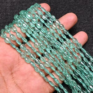 Könnte beinhalten: Mehrere Stränge ovaler, durchscheinender, helltürkisfarbener Perlen. Die Perlen sind aufgefädelt und bilden mehrere Stränge. Die Perlen haben ein glattes, poliertes Aussehen und sind gleichmäßig in Größe und Form. Der Hintergrund ist dunkelgrau.