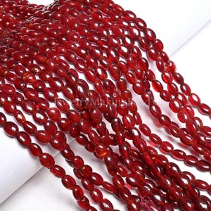 Puede incluir: Hebras de cuentas rojas ovaladas y translúcidas. Las cuentas están ensartadas, creando una apariencia fluida y texturizada. Las cuentas son de un color rojo intenso y rico, con un acabado brillante. La imagen está bien iluminada, resaltando los detalles de las cuentas.