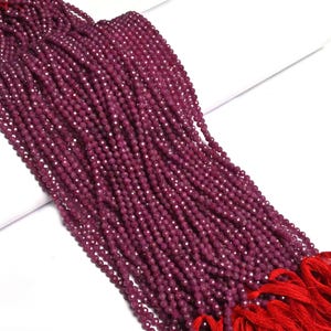 May include: A long strand of small, faceted, deep magenta-colored beads arranged in multiple parallel rows. The beads are strung together and finished with bright red thread tassels at the bottom, creating a uniform and textured appearance.