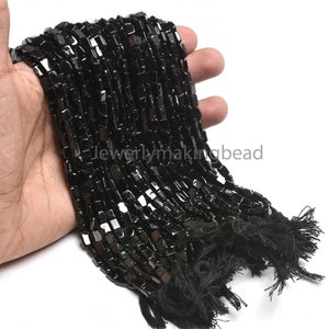 Puede incluir: Un hilo de cuentas cuadradas negras brillantes. Las cuentas se sostienen en una mano, con borlas negras en los extremos del hilo. Las cuentas son uniformes en tamaño y forma, reflejando la luz.