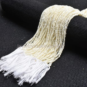 Könnte beinhalten: Kleine, runde, cremefarbene Perlen, die zu mehreren Strängen aufgereiht sind. Die Stränge sind unten mit weißen Quasten zusammengefasst. Die Perlen werden auf einer dunklen, strukturierten Oberfläche präsentiert.