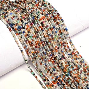 Può includere: Primo piano di più fili di piccole perline di pietre preziose sfaccettate. Le perline sono in una varietà di colori, tra cui blu, arancione, verde e marrone. Sono infilate insieme, creando una superficie strutturata.