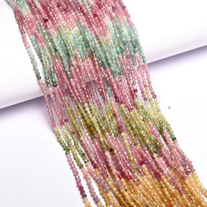 Peut inclure: Gros plan de plusieurs brins de petites perles à facettes dans des tons de rose, de vert et de jaune. Les perles sont enfilées ensemble, créant un effet de cascade. Les perles semblent être des pierres précieuses, peut-être de la tourmaline, sur fond blanc.