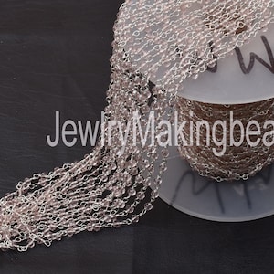 Puede incluir: Un carrete de cadena plateada con cuentas de vidrio rosa claro. La cadena está envuelta alrededor de un carrete blanco con el texto "JewelryMakingbea" impreso en él.