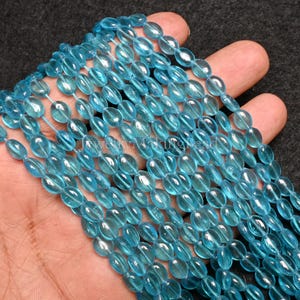 May include: Strands of light blue oval-shaped beads. The beads are strung together, creating a flowing, textured appearance. The beads are a translucent, light blue color. The image is of a hand holding the strands of beads.