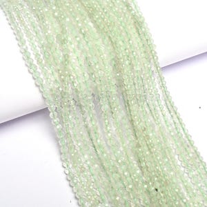 Puede incluir: Hebras de cuentas facetadas de color verde pálido. Las pequeñas cuentas redondas están ensartadas para crear una apariencia fluida y texturizada. Las cuentas tienen un brillo sutil, que refleja la luz. El fondo es blanco.